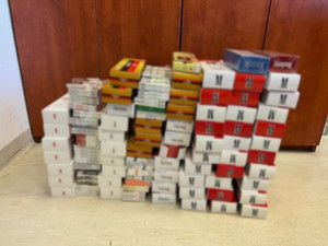 Zdjęcie przedstawia paczki papierosów różnych marek ułożone na podłodze przed brązową szafą.