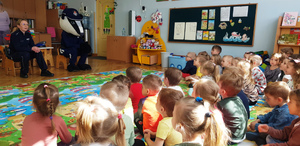 Zdjęcie przedstawia umundurowaną policjantkę, która siedzi obok osoby przebranej za maskotkę Borsuka. Przed nimi na dywanie siedzą dzieci i ich opiekunki z przedszkola.