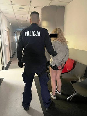 Zdjęcie przedstawia umundurowanego policjanta, który prowadzi przez korytarz zatrzymaną 20-latkę. Oboje stoją tyłem do zdjęcia. Kobieta ma twarz zasłoniętą czarnym prostokątem. Na rękach trzymanych z tyłu ma założone kajdanki. Ubrana jest w szarą bluzę dresową, krótkie spodenki i jasne buty sportowe.