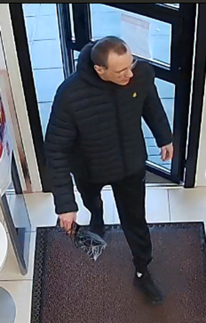 Zdjęcie przedstawia wchodzącego do apteki mężczyznę. Jest on w wieku około 35 lat, wysoki, ma szczupłą budowę ciała, ciemne włosy ostrzyżone na krótko, okulary korekcyjne; ubrany jest w czarną zimową kurtkę z białym symbolem po lewej stronie klatki piersiowej, czarne spodnie dresowe i czarne sportowe buty. W prawej dłoni trzyma ciemną zwiniętą reklamówkę.