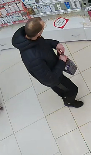 Zdjęcie przedstawia mężczyznę stojącego przed półką z asortymentem. Jest on w wieku około 35 lat, wysoki, ma szczupłą budowę ciała, ciemne włosy ostrzyżone na krótko, okulary korekcyjne; ubrany jest w czarną zimową kurtkę, czarne spodnie dresowe i czarne sportowe buty. W dłoniach trzyma ciemną reklamówkę. Stoi prawym profilem do kamery monitoringu.