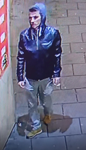 Zdjęcie przedstawia mężczyznę w wieku około 40 lat ubranego w ciemną kurtkę, jasne spodnie, który stoi na zewnątrz obok ściany z plakatem.