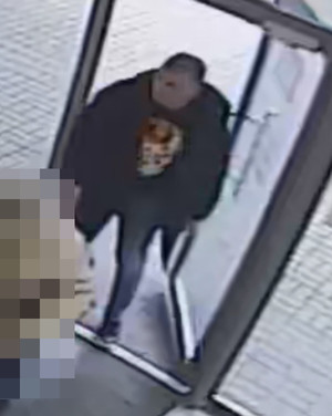 Zdjęcie przedstawia sylwetkę mężczyzny wchodzącego d pomieszczenia z przeszklonymi drzwiami. Mężczyzna jest ubrany w ciemną kurtkę, ciemną bluzę z czerwonym elementem na klatce piersiowej i ciemne spodnie.