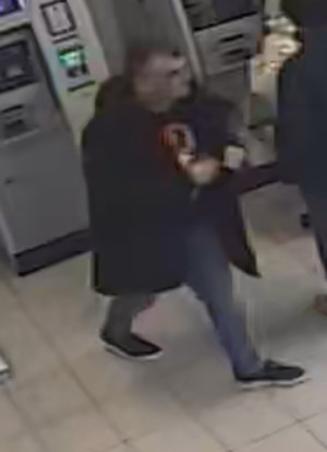 Zdjęcie przedstawia sylwetkę mężczyzny odchodzącego od bankomatu. Mężczyzna jest ubrany w ciemną kurtkę, ciemną bluzę z czerwonym elementem na klatce piersiowej, ciemne spodnie i ciemne buty. W tle widać fragment sylwetki innej osoby, ubranej na ciemno i stojącej przy bankomacie, tyłem do kamery.
