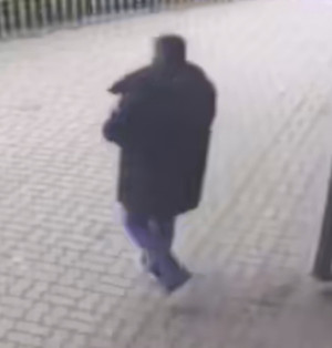 Zdjęcie przedstawia sylwetkę mężczyzny ubranego w ciemną kurtkę, ciemne spodnie i ciemne buty. W tle widać fragment sylwetki innej osoby, ubranej na ciemno i stojącej przy bankomacie, tyłem do kamery.