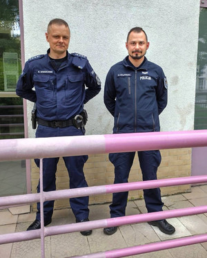 Zdjęcie przedstawia dwóch umundurowanych policjantów, którzy stoją obok siebie przed budynkiem i patrzą wprost do obiektywu aparatu.