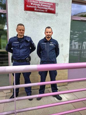 Zdjęcie przedstawia dwóch umundurowanych policjantów, którzy stoją obok siebie przed budynkiem i patrzą wprost do obiektywu aparatu. Nad ich głowami, na ścianie budynku umieszczona jest czerwona tablica z białym napisem: Ii II Rewir Dzielnicowych Wydziału Prewencji Komendy Rejonowej Policji Warszawa VI&amp;quot;.