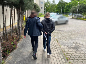 Zdjęcie przedstawia policjanta ubranego w cywilną odzież, który prowadzi po dziedzińcu, po swojej prawej stronie zatrzymanego mężczyznę. Mężczyzna ten jest ubrany na ciemno. Na rękach trzymanych z tyłu ma założone kajdanki. W tle widać zaparkowany oznakowany radiowóz.