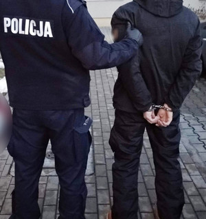 Zdjęcie przedstawia umundurowanego policjanta, który prowadzi po swojej prawej stronie zatrzymanego mężczyznę. Mężczyzna ten ma kajdanki założone na rekach trzymanych z tyłu. Obaj stoją tyłem do obiektywu aparatu.