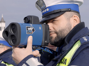 Zdjęcie przedstawia umundurowanego policjanta ruchu drogowego w białej czapce, który patrzy w ekran miernika prędkości.