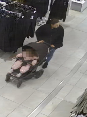Zdjęcie przedstawia kobietę wewnątrz sklepu. Rysopis kobiety: wiek około 20 lat, szczupła budowa ciała, włosy w kolorze ciemnym upięte w kucyk; ubrana jest w czarną kurtkę, spodnie jeansowe w kolorze jasnoniebieskim, czarne buty sportowe. Przed sobą prowadzi ciemny wózek tzw. &quot;spacerówkę&quot; z dzieckiem w środku.