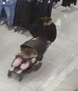 Zdjęcie przedstawia kobietę wewnątrz sklepu. Rysopis kobiety: wiek około 20 lat, szczupła budowa ciała, włosy w kolorze ciemnym upięte w kucyk; ubrana jest w czarną kurtkę, spodnie jeansowe w kolorze jasnoniebieskim, czarne buty sportowe. Przed sobą prowadzi ciemny wózek tzw. &quot;spacerówkę&quot; z dzieckiem w środku.