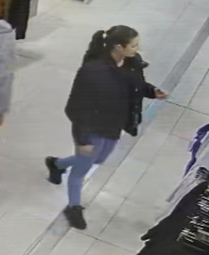 Zdjęcie przedstawia kobietę wewnątrz sklepu - wiek około 20 lat, szczupła budowa ciała, włosy w kolorze ciemnym upięte w kucyk; ubrana jest w czarną kurtkę, spodnie jeansowe w kolorze jasnoniebieskim, czarne buty sportowe. Kobieta znajduje się prawym profilem do kamery.