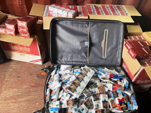 Zdjęcie przedstawia otwarte brązowe kartony i otwartą czarną walizkę zawierające paczki papierosów.