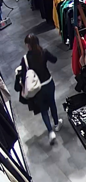 Zdjęcie przedstawia kobietę w sklepie - wiek około 28 lat, czarne związane włosy; ubrana jest w czarną skórzaną kurtkę, niebieskie spodnie jeansowe oraz białe sportowe buty, na ramieniu nosi jasną torbę. Stoi tyłem do kamery monitoringu.