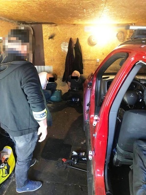 Zdjęcie przedstawia mężczyznę stojącego tyłem do obiektywu aparatu, który ma odblaskową opaskę z napisem POLICJA na prawym przedramieniu. Mężczyzna stoi obok czerwonego pojazdu zaparkowanego tyłem. Przed nim widać dwie osoby siedzące na ziemi w pomieszczeniu.