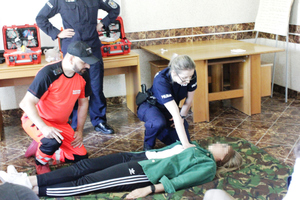 Zdjęcie przedstawia policjanta ubranego w mundur i stojącego za dwoma osobami klęczącymi przed leżącą na podłodze kobietą. Kobieta kucająca jet ubrana w granatowy mundur. Mężczyzna obok niej w odblaskowy pomarańczowy uniform ratownika medycznego.
