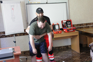 Zdjęcie przedstawia mężczyznę ubranego w odblaskowy pomarańczowy uniform ratownika medycznego, który obejmuje rękami stojącego przed nim mężczyznę ze zwieszonymi rekami.