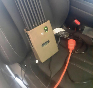 Zdjęcie przedstawia ułożone na fotelu wewnątrz pojazdu urządzenie elektroniczne, od którego odchodzi czarny i czerwony kabel.