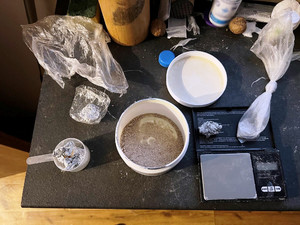 Zdjęcie przedstawia stół, na którym znajdują się pojemniki z różnymi substancjami, woreczki foliowe z jakąś zawartością, czarna waga elektroniczna i zawiniątka z folii aluminiowej.