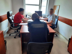 Zdjęcie przedstawia dwóch nieumundurowanych policjantów, którzy siedzą po obu stronach biurka, naprzeciwko siebie przed monitorami komputera. Obaj mają zasłonięte twarze. Jeden ma na sobie czerwoną koszulkę na krotki rękaw, drugi białą koszulę. Zatrzymany mężczyzna siedzi przy biurku naprzeciwko nich. Jest ubrany w ciemną bluzę. Siedzi tyłem do wejścia do pokoju. Napis na jego plecak jest zasłonięty.