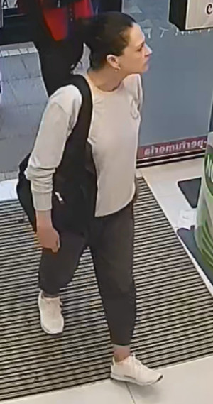 Zdjęcie przedstawia kobietę wchodzącą do drogerii. Jest ona w wieku około 30-40 lat, ma szczupłą budowę ciała, włosy w kolorze czarnym, spięte w kucyk; ubrana jest w szarą bluzę, czarne spodnie, białe buty, miała ze sobą czarny plecak.