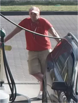 Zdjęcie przedstawia mężczyznę stojącego pomiędzy dystrybutorem paliwa i ciemnym samochodem. Mężczyzna jest w wieku około 40-50 lat, ubrany w jasne krótkie spodnie, czerwony t-shirt, czapkę z daszkiem, okulary przeciwsłoneczne.