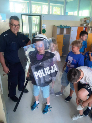 Zdjęcie przedstawia dzieci stojące obok umundurowanego policjanta. Jeden z chłopców trzyma przed sobą tarczę z napisem POLICJA, w prawe dłoni pałkę policyjną, zaś na głowie ma policyjny kask.