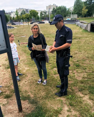 Zdjęcie przedstawia umundurowanego policjanta, który stoi przy kobiecie na placu zabaw. Obok niej widać dziewczynkę. Kobieta trzyma w dłoniach broszurę informacyjną.