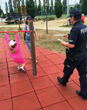 Zdjęcie przedstawia umundurowanego policjanta, który podaje ulotkę edukacyjną kobiecie. Obok niej widać kilkuletnią dziewczynkę, która zwisa z drążka.