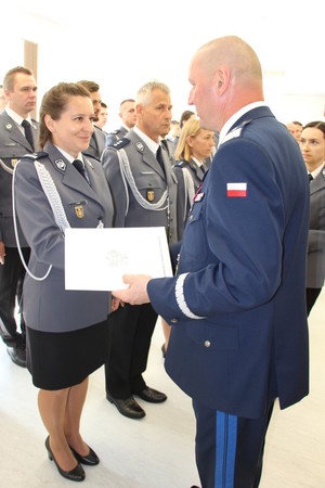 Zdjęcie przedstawia policjanta w granatowym mundurze, który stoi naprzeciwko policjantki w granatowym mundurze i podaje jej rękę. W lewej dłoni trzyma jasną teczkę.