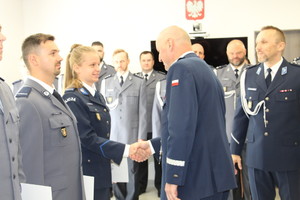 Zdjęcie przedstawia policjanta w granatowym mundurze, który stoi naprzeciwko policjantki w takim samym granatowym mundurze i podaje jej rękę. W lewej dłoni trzyma jasną teczkę.