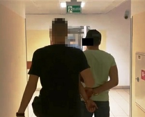 Zdjęcie przedstawia sylwetki dwóch osób idących korytarzem. Osoba z lewej strony to policjant ubrany w ciemną koszulkę na krótki rękaw. Twarz policjanta jest zasłonięta komputerowym filtrem, po jego prawej stronie idzie zatrzymany mężczyzna. Ma na sobie koszulkę na krótki rękaw, w jasnym kolorze i czarną czapkę. Część twarzy zasłania mu czarny prostokąt. Na rękach trzymanych z tyłu ma założone kajdanki. Obaj stoją tyłem do obiektywu aparatu.