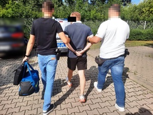 Zdjęcie przedstawia dwóch policjantów ubranych w cywilną odzież, którzy prowadzą pomiędzy sobą zatrzymanego mężczyznę. Policjanci mają zasłonięte twarze komputerowym filtrem. Mężczyzna prowadzony pomiędzy nimi ma kajdanki założone na rekach trzymanych z tyłu. Policjant idący po lewe stronie trzyma w lewej ręce niebieską torbę sportową. Przed nimi widać tył zaparkowanego radiowozu.