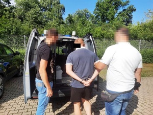 Zdjęcie przedstawia dwóch policjantów ubranych w cywilną odzież, którzy stoją z zatrzymanym mężczyzną. Policjanci mają zasłonięte twarze komputerowym filtrem. Mężczyzna prowadzony pomiędzy nimi ma kajdanki założone na rekach trzymanych z tyłu. Policjant stojący po lewej stronie otwiera drzwi do zaparkowanego radiowozu.