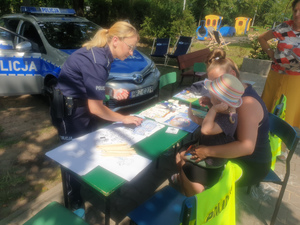 Zdjęcie przedstawia umundurowaną policjantkę, która pochyla się nad stołem, przy którym siedzi mała dziewczynka w kapeluszu na kolanach babci. Na stole widać rozłożone materiały. Za nią widać oznakowany radiowóz.