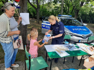 Zdjęcie przedstawia umundurowaną policjantkę, która pochyla się nad małą dziewczynką i wręcza jej kolorowankę. Na stole widać rozłożone materiały. Za nimi stoi oznakowany radiowóz.