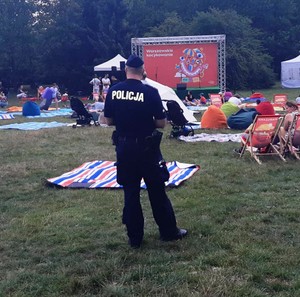 Zdjęcie przedstawia umundurowanego policjanta, który stoi w przestrzeni publicznej w parku. Przed nim widać leżaki osoby przebywające w parku.