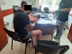 Zdjęcie przedstawia mężczyznę siedzącego przy stole. Po jego prawej stronie widać stojącego policjanta. Mężczyzna ma kajdanki na rękach. Połowę jego twarzy zasłania czarny prostokąt.