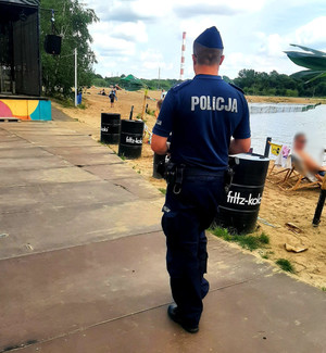 Zdjęcie przedstawia sylwetkę umundurowanego policjanta, który idzie deptakiem wzdłuż plaży.