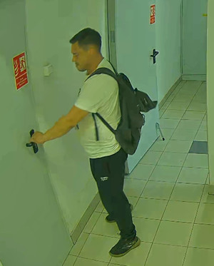 Zdjęcie przedstawia mężczyznę stojącego na korytarzu. Mężczyzna ten jest skierowany lewym profilem do kamery. Chwyta za klamkę drzwi. Mężczyzna ma około 30 lat, około 180 cm wzrostu, grubszą posturę ciała; ubrany jest w białą koszulkę, czarne spodnie, czarne sportowe buty, ma czarny plecak.