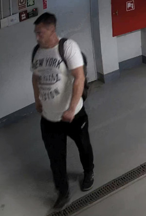 Zdjęcie przedstawia mężczyznę idącego korytarzem. Mężczyzna ten jest skierowany lewym profilem do kamery. Ma około 30 lat, około 180 cm wzrostu, grubszą posturę ciała; ubrany jest w białą koszulkę, czarne spodnie, czarne sportowe buty, ma czarny plecak. Dłonie trzyma w kieszeniach spodni.