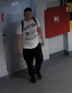 Zdjęcie przedstawia mężczyznę na korytarzu. Ma około 30 lat, około 180 cm wzrostu, grubszą posturę ciała; ubrany jest w białą koszulkę z napisem: „NEW YORK DIVISION”, czarne spodnie, czarne sportowe buty, ma plecak.