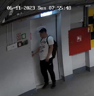 Zdjęcie przedstawia mężczyznę na korytarzu. Ma około 30 lat, około 180 cm wzrostu, grubszą posturę ciała; ubrany jest w białą koszulkę z napisem: „NEW YORK DIVISION”, czarne spodnie, czarne sportowe buty, ma plecak.