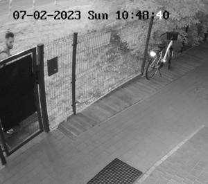 Zdjęcie przedstawia działkę przed domem. Za ogrodzeniem, w lewym rogu zdjęcia widać głowę mężczyzny. Zdjęcie jest w odcieniach szarości.