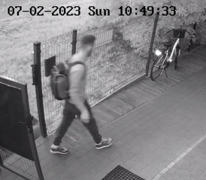 Zdjęcie przedstawia mężczyznę wchodzącego na działkę przez otwartą furtkę. Na działce, przy ogrodzeniu stoi rower, po prawej stronie zdjęcia. Zdjęcie jest w odcieniach szarości.