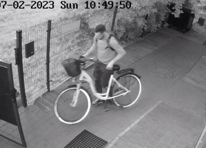 Zdjęcie przedstawia mężczyznę wyprowadzającego rower chodzącego z działki. Zdjęcie jest w odcieniach szarości.