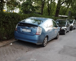 Zdjęcie przedstawia trzy samochody osobowe zaparkowane jeden za drugim. Dwa z nich są w kolorze ciemnym, ostatni w kolorze niebieskim. Ma on zasłonięte tablice rejestracyjne. Za pojazdami widać zielone zarośla i drzewa.