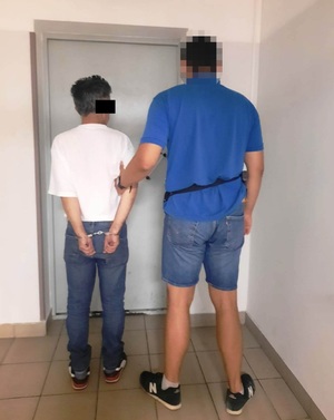 Zdjęcie przedstawia policjanta ubranego w niebieską koszulkę na krótki rękaw i granatowe spodenki jeansowe, który prowadzi po swojej lewej stronie zatrzymanego mężczyznę.