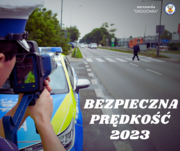 Zdjęcie przedstawia fragment sylwetki umundurowanego policjanta, który patrzy w obiektyw radaru na pojazdy znajdujące się na pasie ruchu. Przed nim, w tle widać oznakowany radiowóz.